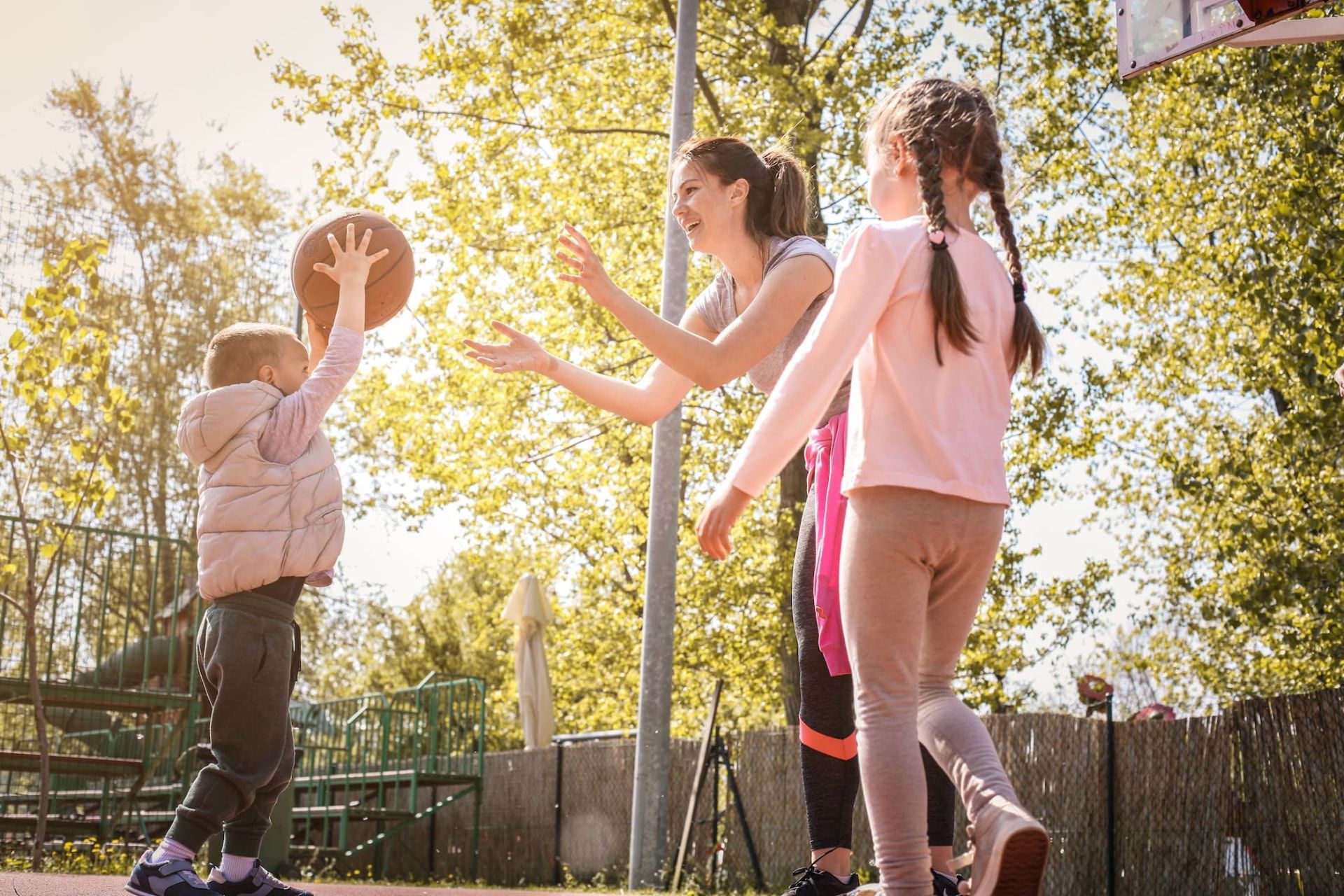 A nanny playing basketball outdoors with a toddler and an older child